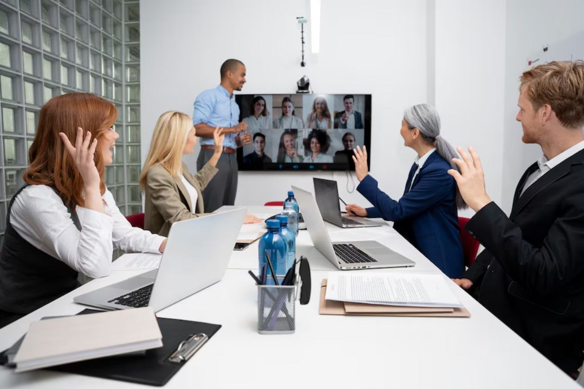 A group of professionals in a conference room engaged in a virtual meeting, with laptops and documents on the table.
