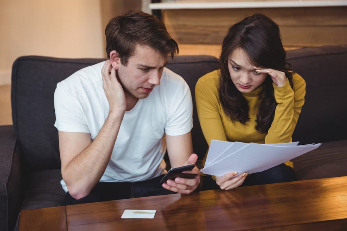 A man in a white T-shirt and a woman in a yellow sweater review documents together on a couch, looking concerned.