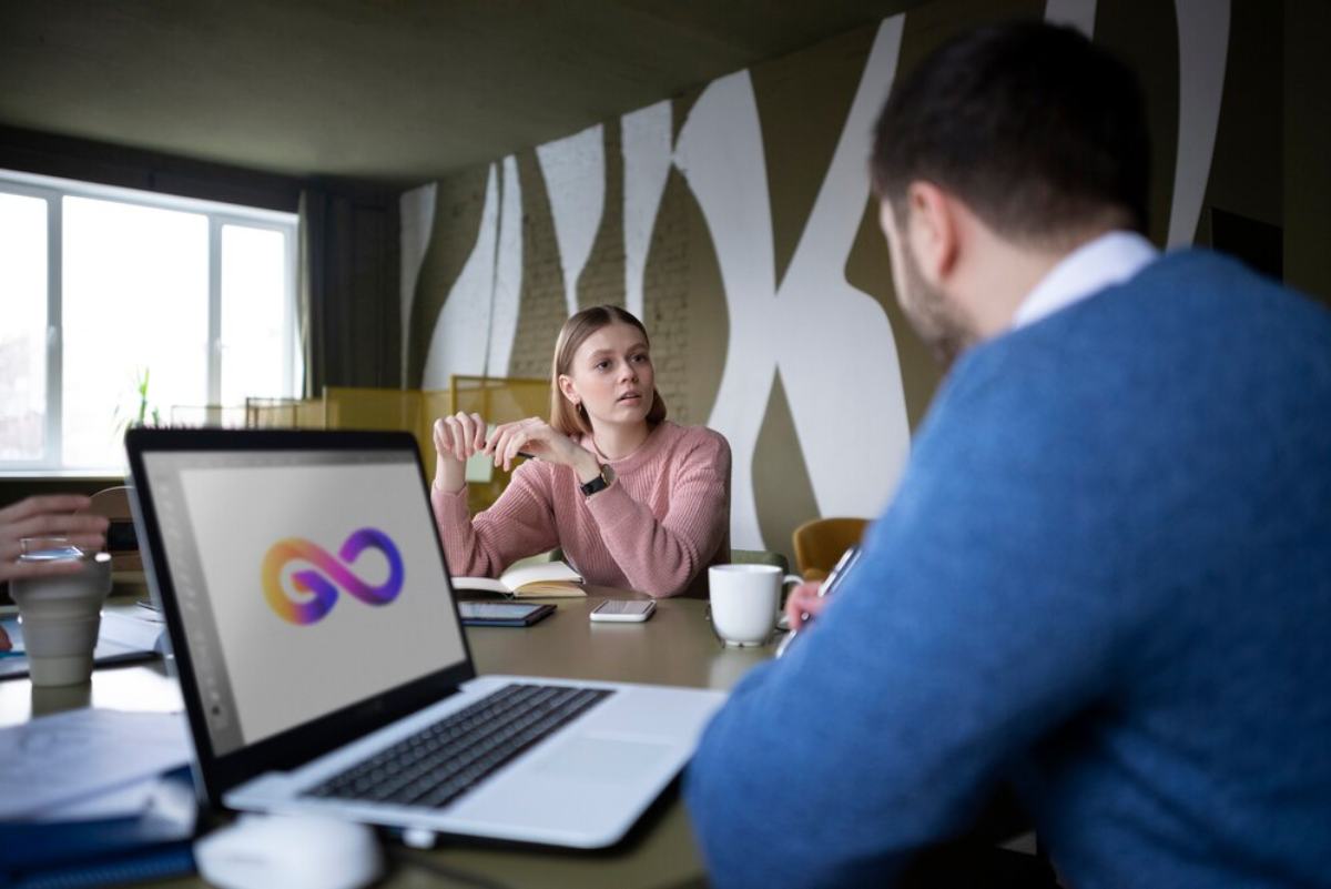 A laptop displays a colorful infinity logo during a meeting, with two people engaged in conversation at a stylish, modern workspace.