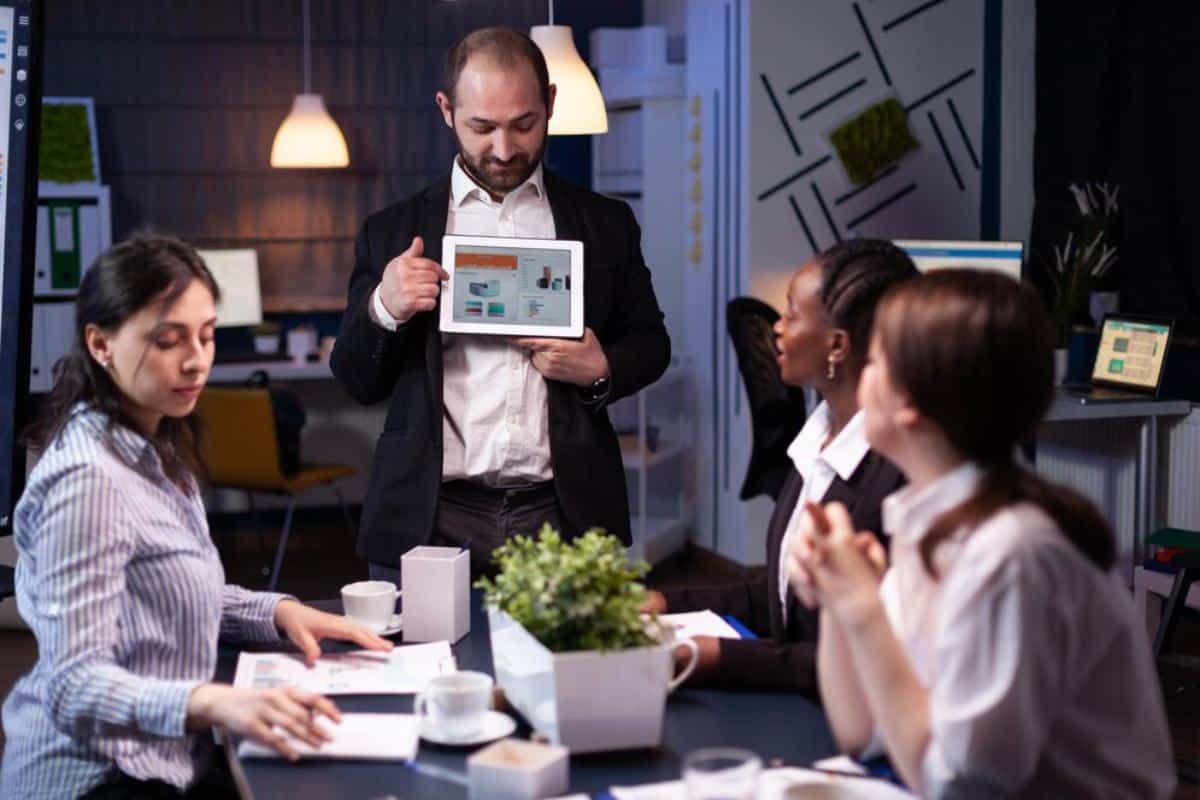 A group of professionals engage in a meeting, with one person presenting data on a tablet while others listen attentively.