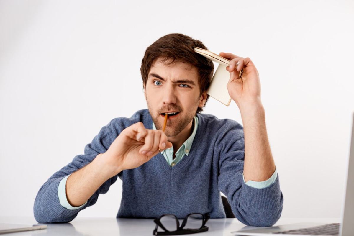 A person in a blue sweater sits at a desk, holding a notebook to their head with a thoughtful expression, surrounded by a laptop and glasses.