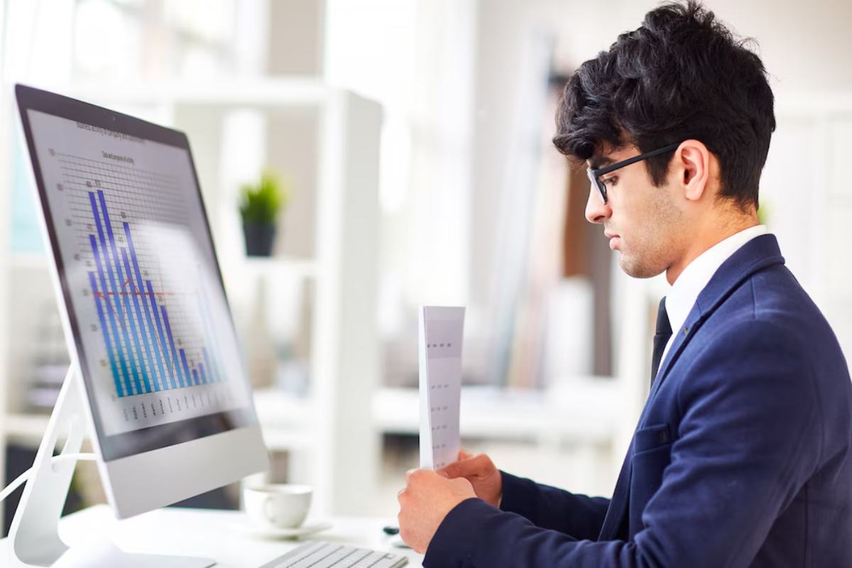 A man in a suit analyzes a graph on a computer screen while holding a printed document in a bright, modern office setting.