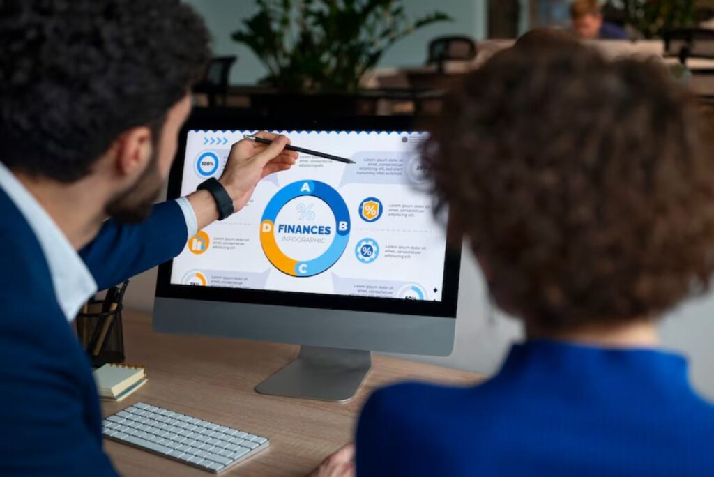 A person points at a finance infographic on a computer screen, while another individual observes, in a modern office setting.