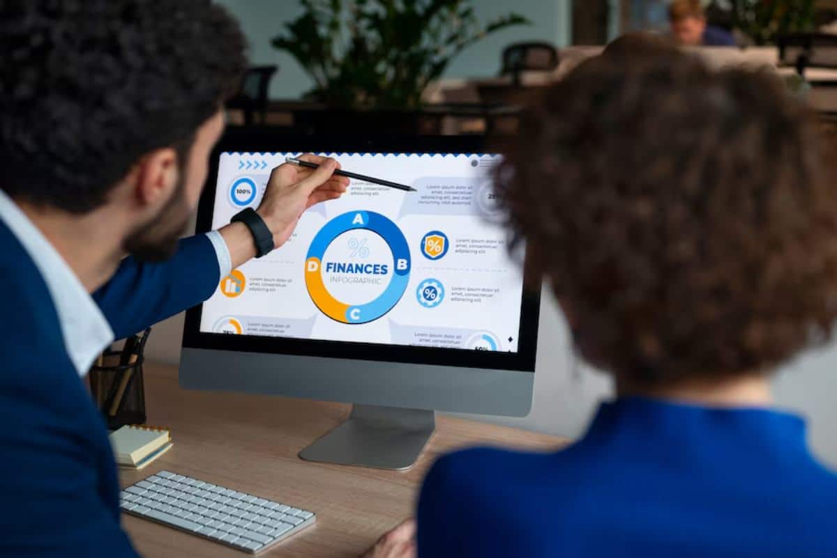 A person points at a finance infographic on a computer screen, while another individual observes, in a modern office setting.