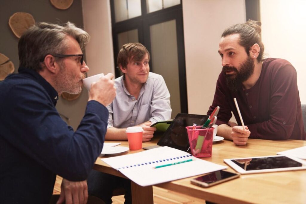 A group of three people engage in a discussion at a wooden table, surrounded by notebooks, tablets, and cups. Collaboration in a cozy setting.