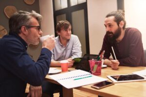 A group of three people engage in a discussion at a wooden table, surrounded by notebooks, tablets, and cups. Collaboration in a cozy setting.