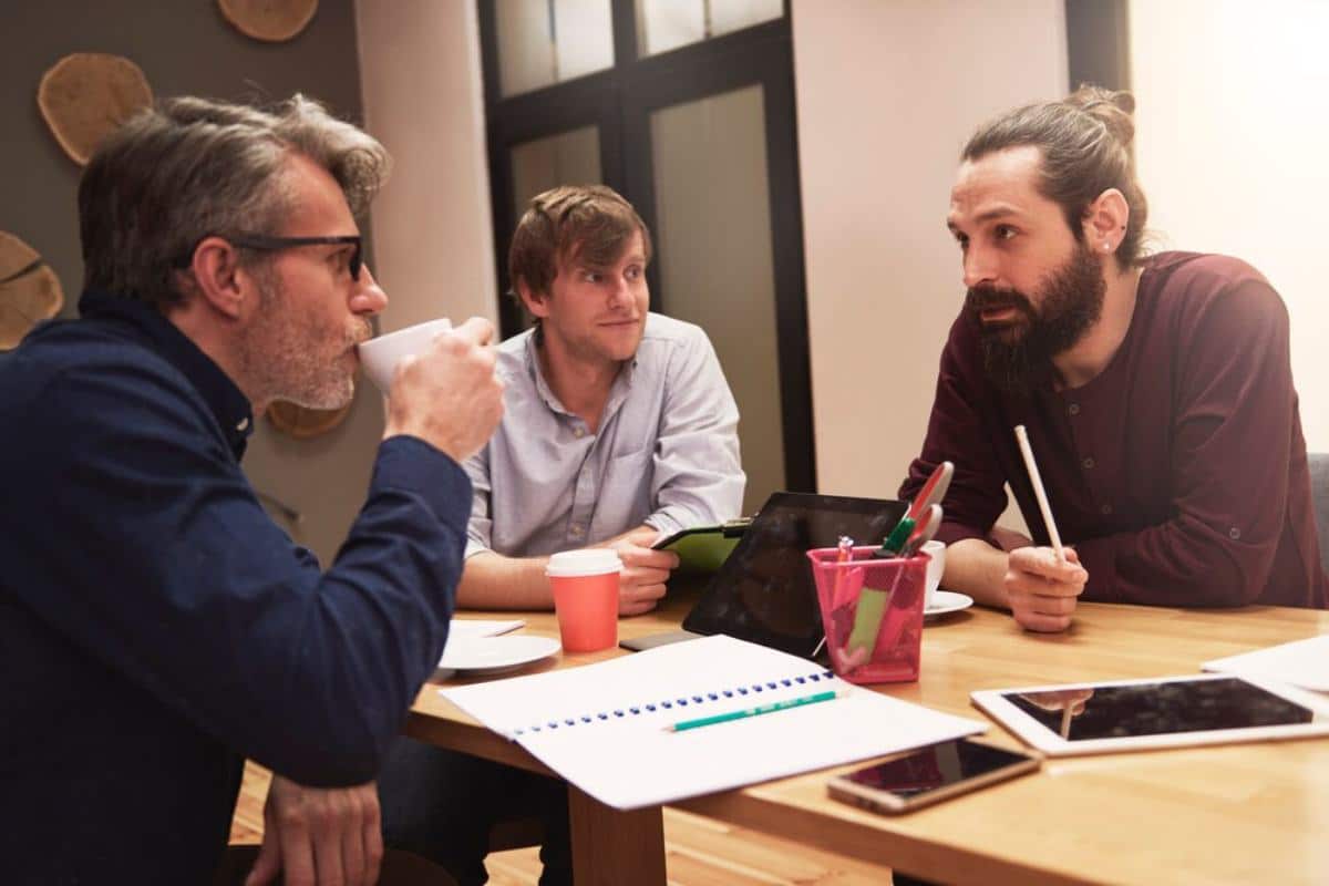 A group of three people engage in a discussion at a wooden table, surrounded by notebooks, tablets, and cups. Collaboration in a cozy setting.