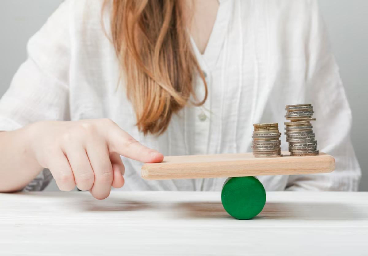 A woman's hand balances a wooden plank with stacks of coins on one side, symbolizing finance and equilibrium.